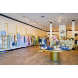 The boutique interior at Primm's showcases a range of colorful garments beautifully arranged on clothing racks along the walls. A round table in the foreground features neatly folded clothes and stylish accessories. The wooden flooring and decorative plants create a cozy and welcoming ambiance, ideal for a Personal Styling Appointment.