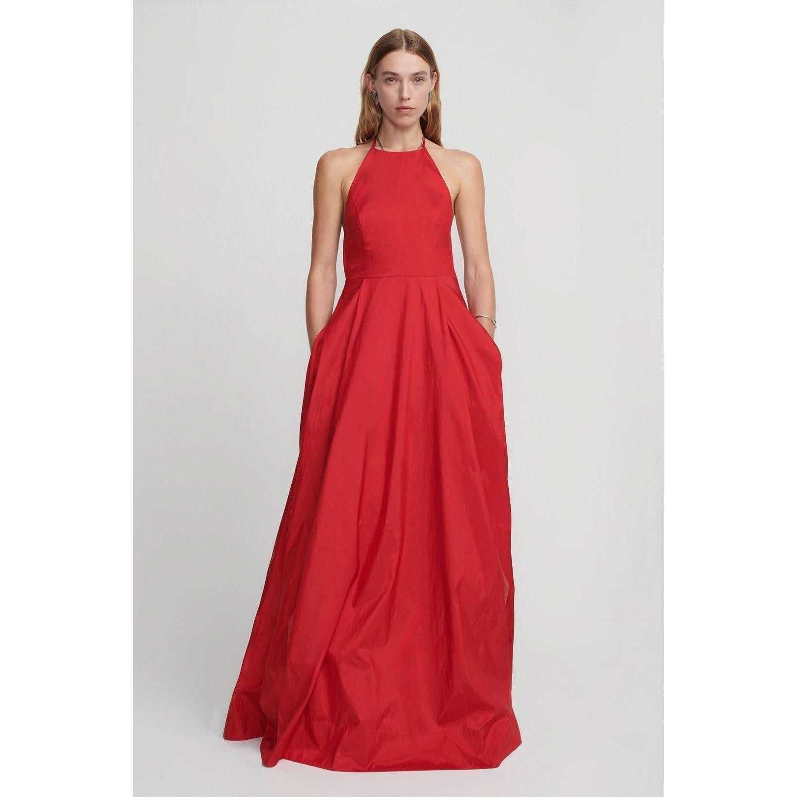 A woman stands against a plain background, wearing the Hansen & Gretel Gilda Gown Samba from Hansen & Gretel. The floor-length red silk maxi dress features a halter neck and a fitted bodice that flares into a full skirt. She has one hand in her pocket and a neutral expression on her face.