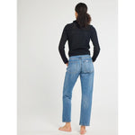 A person with curly hair is standing barefoot, facing away from the camera. They are wearing blue jeans and the Mille Andie Cardigan Black by Mille, featuring a hand crochet trim. The background is plain white.