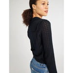 A person with curly hair is wearing a Mille Andie Cardigan Black with hand crochet trim and blue jeans. They are looking over their shoulder with a slight smile, against a plain light-colored background.