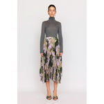A person stands against a plain background wearing a gray turtleneck paired with the Christy Lynn Gwen Skirt - Midnight Garden, a high-waisted, pleated midi skirt featuring a floral pattern. With earrings and sandals adding charm, the look is completed by the fitted waist that subtly highlights their silhouette.