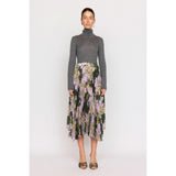 A person stands against a plain background wearing a gray turtleneck paired with the Christy Lynn Gwen Skirt - Midnight Garden, a high-waisted, pleated midi skirt featuring a floral pattern. With earrings and sandals adding charm, the look is completed by the fitted waist that subtly highlights their silhouette.