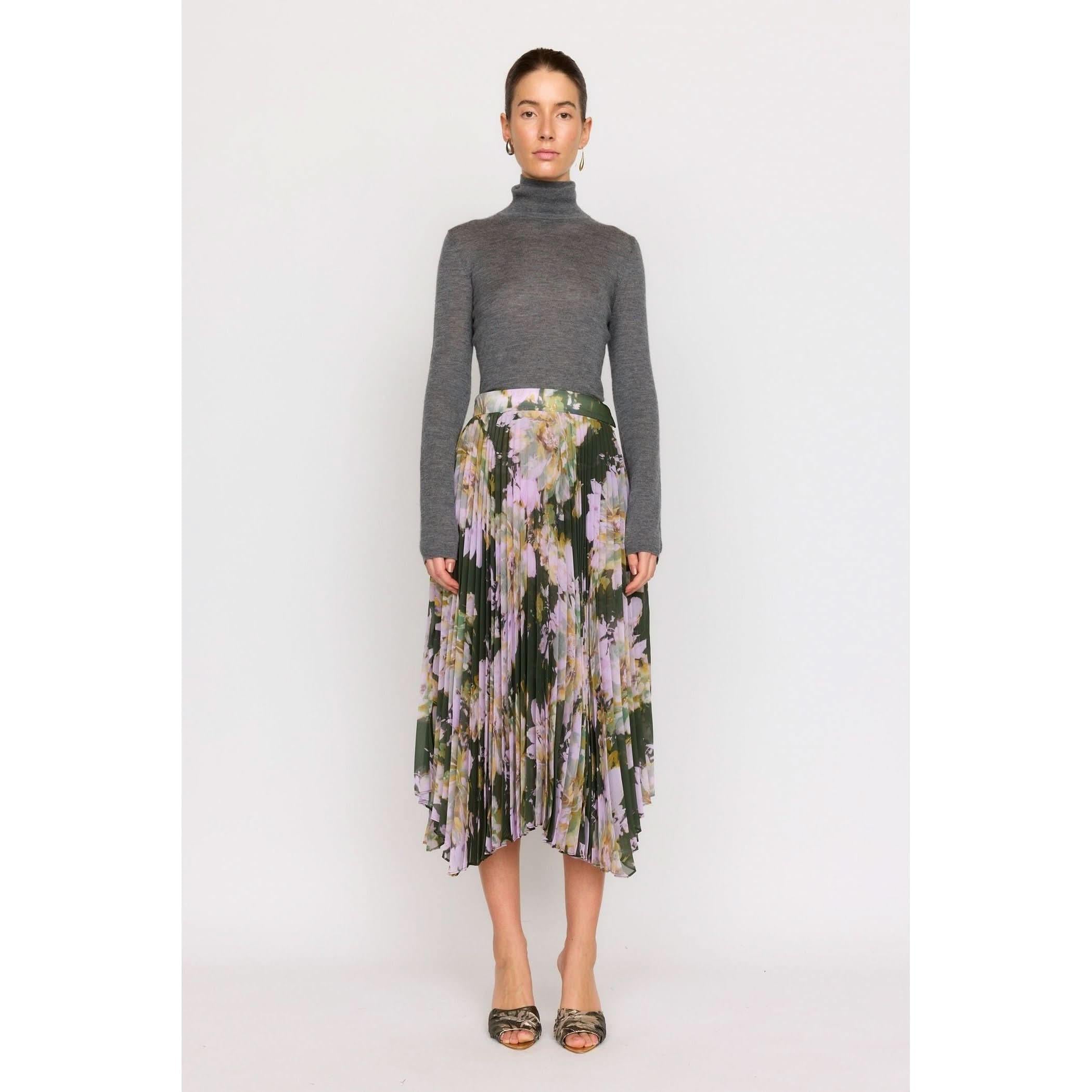 A person stands against a plain background wearing a gray turtleneck paired with the Christy Lynn Gwen Skirt - Midnight Garden, a high-waisted, pleated midi skirt featuring a floral pattern. With earrings and sandals adding charm, the look is completed by the fitted waist that subtly highlights their silhouette.