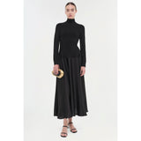 Dressed in a sleek Simkhai Frances Dress, a person stands gracefully holding a small, gold, heart-shaped object. The dress features a high neckline and pintucked waist, while their hair is neatly pulled back and they are wearing black sandals. The simple white background enhances the elegance of this ensemble by Simkhai.