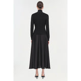 A woman with her hair in a bun faces away from the camera, displaying the elegant Simkhai Frances Dress. The ensemble's long-sleeved black design highlights a pintucked waist and is paired beautifully with black high-heeled sandals against a pristine white backdrop.