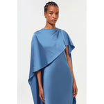 A person stands facing the camera, wearing the Simkhai Izel Cape Gown from the Pre-Fall '24 Lookbook. This elegant gown features draped sleeves and a cape-like design over one shoulder. The background is plain and light-colored.