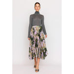 The individual is dressed in a gray turtleneck sweater paired with the Christy Lynn Gwen Skirt in the Midnight Garden design, which showcases a pleated floral pattern with green, pink, and white hues and includes a fitted waist. They are posed against a plain background wearing open-toed shoes.