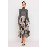 The individual is dressed in a gray turtleneck sweater paired with the Christy Lynn Gwen Skirt in the Midnight Garden design, which showcases a pleated floral pattern with green, pink, and white hues and includes a fitted waist. They are posed against a plain background wearing open-toed shoes.