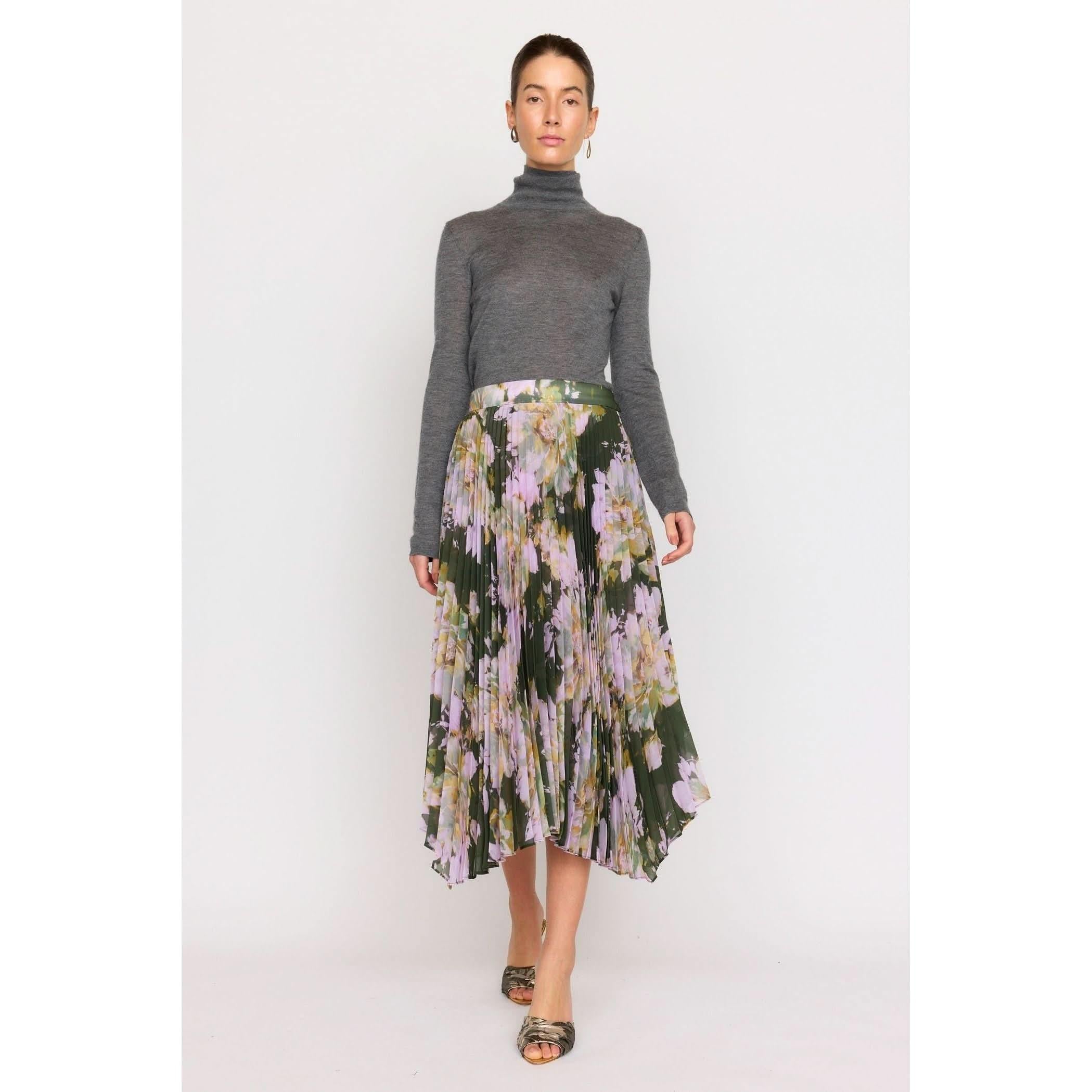 The individual is dressed in a gray turtleneck sweater paired with the Christy Lynn Gwen Skirt in the Midnight Garden design, which showcases a pleated floral pattern with green, pink, and white hues and includes a fitted waist. They are posed against a plain background wearing open-toed shoes.
