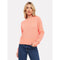 A person wearing the Brodie Cashmere Cathy Cashmere Cable Knit sweater in neon coral, featuring a ribbed crew neckline, stands with one hand in their pocket against a plain white background. They have long, wavy blonde hair and are paired with dark blue jeans.