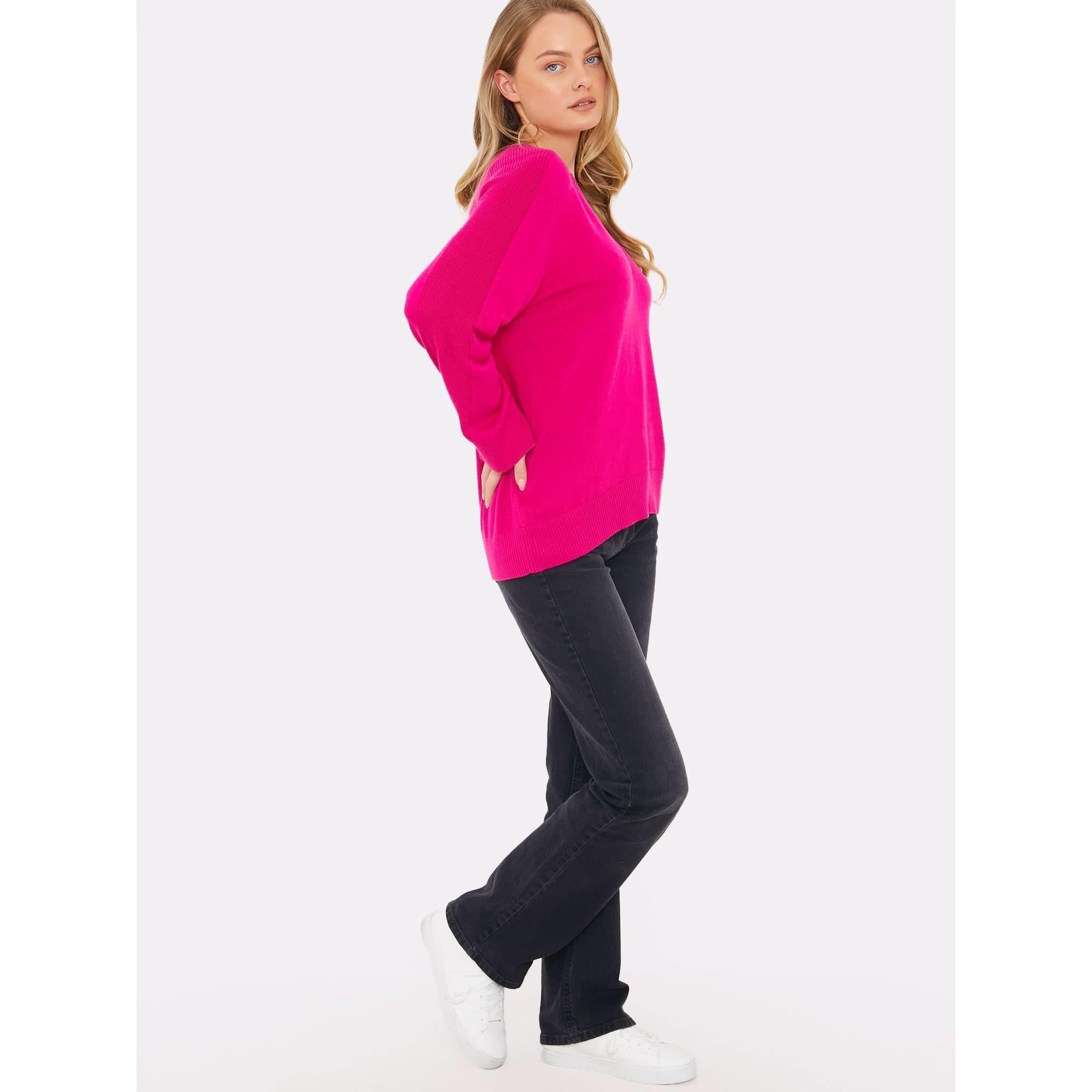 A person with long hair stands sideways, wearing a vibrant fuschia sweater from the Brodie Cashmere collection featuring saddle shoulder rib knit design and a crew neckline, paired with black pants and white sneakers. Their hands are in their pockets, conveying a relaxed expression against a plain white background.