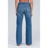 A person is standing facing away from the camera, wearing the ASKK NY Straight Jean Bush Baby. These high-waisted, straight-leg blue jeans are crafted from Japanese denim and feature back pockets with a central seam running down to the hem. The backdrop is plain and light-colored.