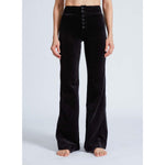 A person stands barefoot on a light background, wearing the ASKK NY Brighton Wide Leg Black Velvet pants. These high-waisted pants feature a button-up front and a sleek finish. The person's hands rest relaxed by their sides.