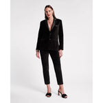 A person stands against a plain background, showcasing the Frances Valentine Tuxedo Jacket in black, featuring a sleek velvet design with a deep neckline. Complementing the look with matching high heels, they have long hair and are adorned with gold bracelets and earrings, presenting a tailored silhouette that exudes elegance.
