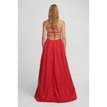 A woman with long, wavy blonde hair is pictured from the back, wearing the Hansen & Gretel Gilda Gown Samba. The strapless red silk maxi dress from Hansen & Gretel features an open back with thin straps tied across her shoulders and a flowing, floor-length skirt. She stands against a plain light gray background.