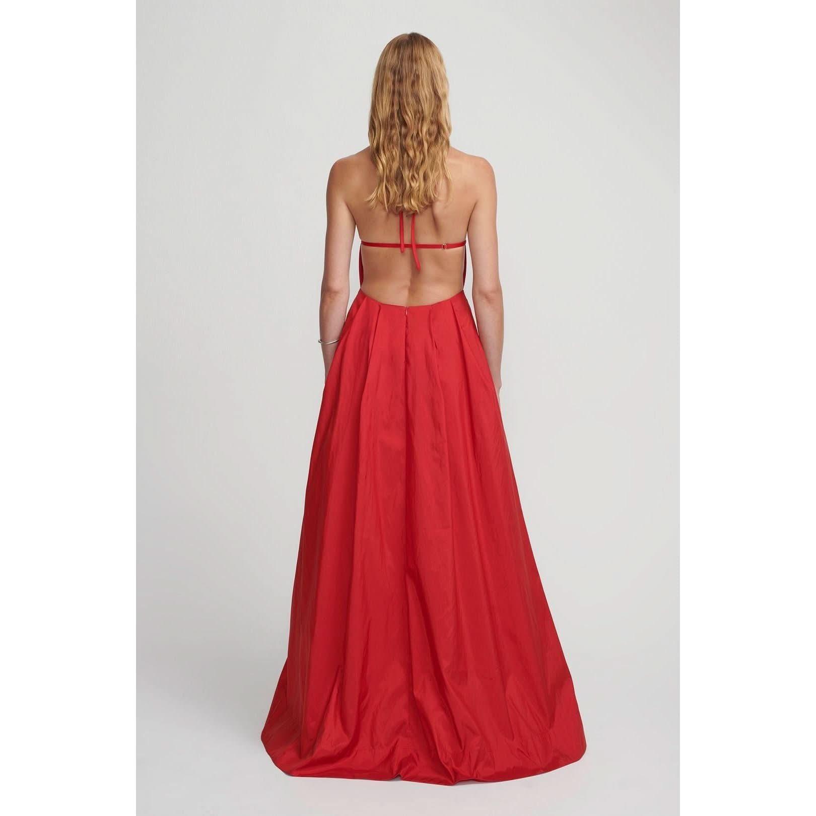 A woman with long, wavy blonde hair is pictured from the back, wearing the Hansen & Gretel Gilda Gown Samba. The strapless red silk maxi dress from Hansen & Gretel features an open back with thin straps tied across her shoulders and a flowing, floor-length skirt. She stands against a plain light gray background.