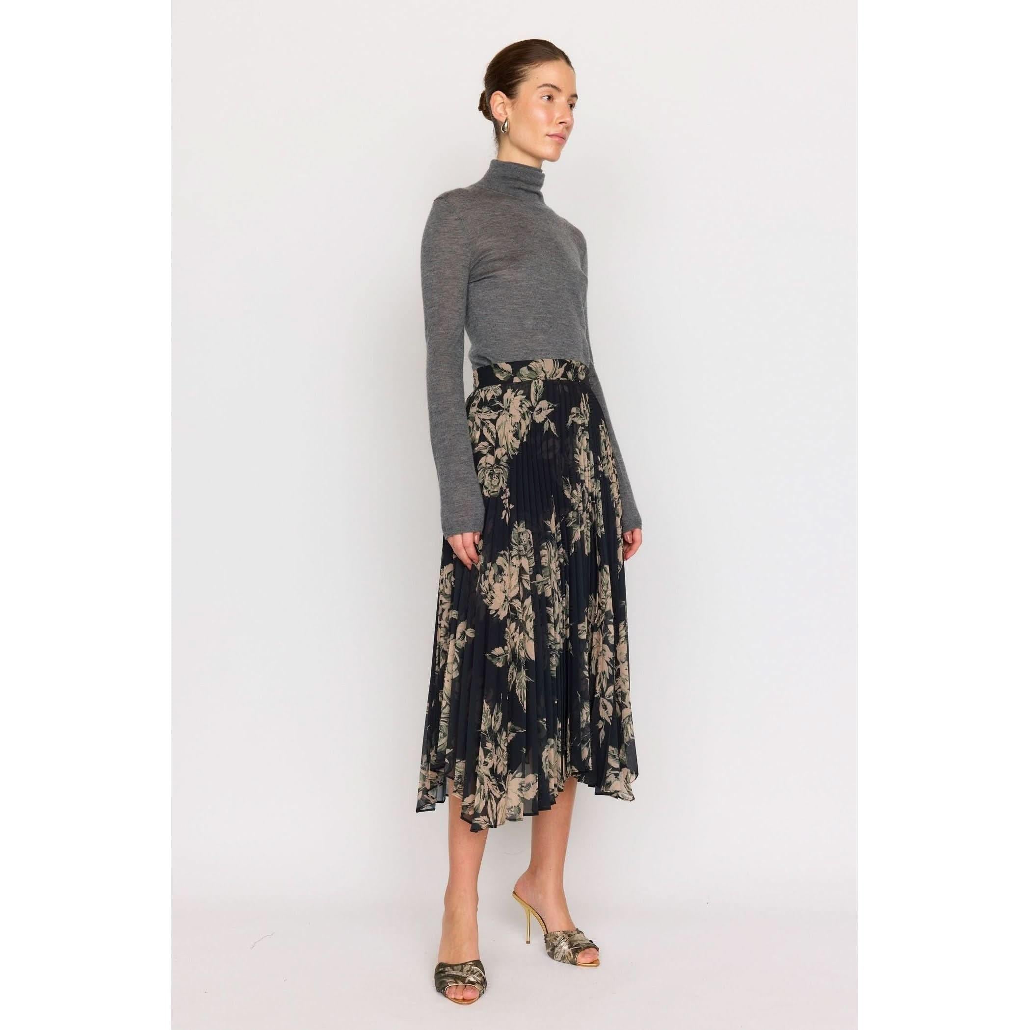 A person wearing a gray turtleneck sweater and the Christy Lynn Gwen Skirt in Black Rose, with its intricate pleating and floral pattern, stands against a plain background. They also sport patterned heels and gold hoop earrings.