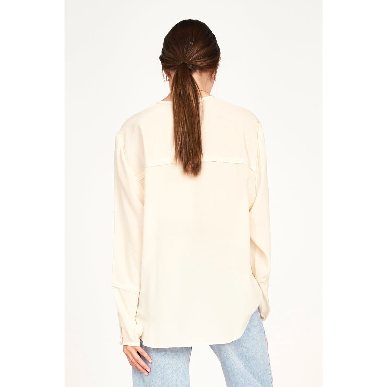 An individual with long brown hair styled in a ponytail is wearing the Mirth Dakota Blouse in Ivory, characterized by its sheer fabric and oversized fit, paired with light blue jeans. They are standing and facing away from the camera against a plain white background.