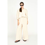 A woman stands confidently, wearing the Mirth Lounge Pant in Dove along with a matching long-sleeve top and oversized sunglasses. The naturally dyed, regenerative cotton outfit complements her dark hair, which is tied back, as she sports black sandals. She poses against a plain white background.