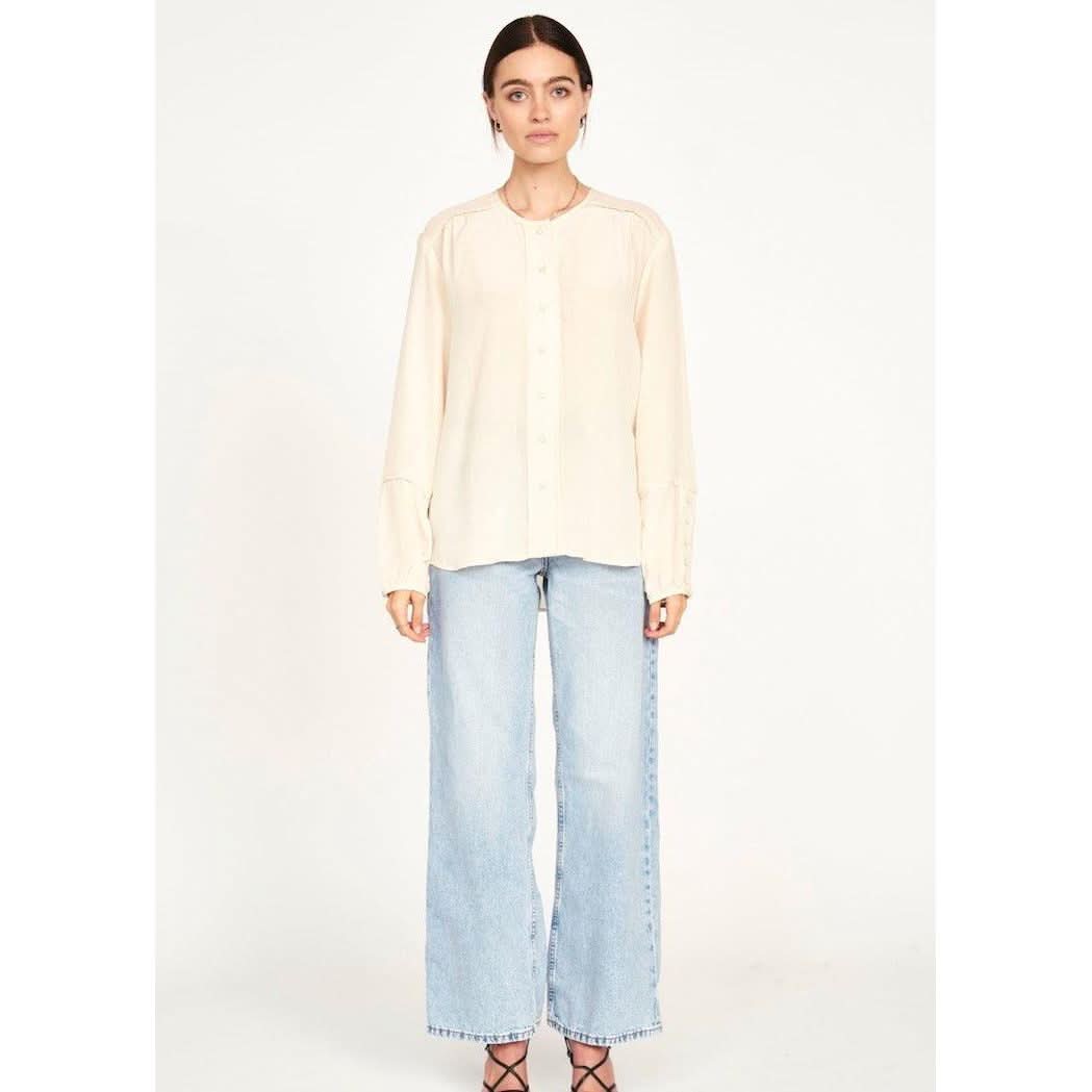 A woman with long dark hair stands against a plain white background. She is wearing an oversized-fit, long-sleeve blouse named the "Mirth Dakota Blouse Ivory" by Mirth and light blue wide-leg jeans. She is also wearing strappy black heels. Her expression is neutral as she faces the camera directly.