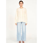 A woman with long dark hair stands against a plain white background. She is wearing an oversized-fit, long-sleeve blouse named the "Mirth Dakota Blouse Ivory" by Mirth and light blue wide-leg jeans. She is also wearing strappy black heels. Her expression is neutral as she faces the camera directly.