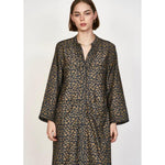 A person with shoulder-length hair is wearing a long-sleeved, patterned Mirth Malmo Dress in Navy Lilypad, made in India. The hand screen-printed design features an intricate, repeating pattern in a mix of brown and dark colors. They are standing against a plain background.