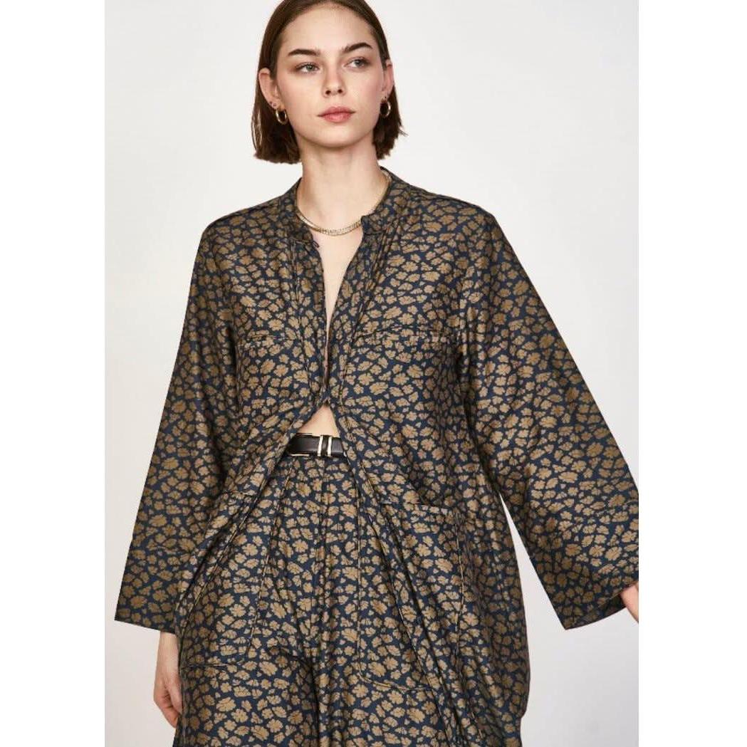 A woman with short brown hair is wearing the Mirth Malmo Dress in Navy Lilypad, which features a dark intricate gold pattern. The hand screen-printed dress, made in India, has an open front that reveals part of her torso. She accessorizes with understated jewelry.