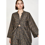 A woman with short brown hair is wearing the Mirth Malmo Dress in Navy Lilypad, which features a dark intricate gold pattern. The hand screen-printed dress, made in India, has an open front that reveals part of her torso. She accessorizes with understated jewelry.