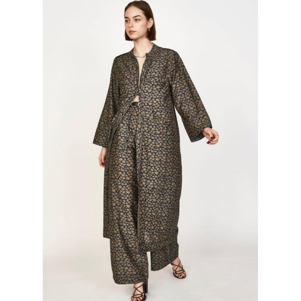 A model wearing the Mirth Malmo Dress in Navy Lilypad, featuring a long, patterned open-front coat and wide-leg trousers. The fabric, hand screen printed and Made in India, showcases a detailed blue and brown geometric design. The model is posing against a plain white background, accessorized with dark strappy sandals.