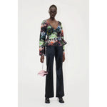 A person stands against a white background, wearing the Stine Goya Nathalie Top Glitter Bloom—a multicolored floral-patterned top with a deep V-neck and peplum detail, crafted from sustainably sourced materials. They pair it with black high-waisted pants and black shoes. In their right hand, they hold a small rectangular light pink handbag with a silver chain.