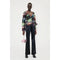 A person stands against a white background, wearing the Stine Goya Nathalie Top Glitter Bloom—a multicolored floral-patterned top with a deep V-neck and peplum detail, crafted from sustainably sourced materials. They pair it with black high-waisted pants and black shoes. In their right hand, they hold a small rectangular light pink handbag with a silver chain.