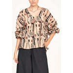 A woman wears the Mirth Petaluma Blouse in Sand Labyrinth, designed with a geometric pattern and crafted from a cotton/silk blend in brown and black tones. Made in India, she pairs the blouse with loose black pants, rolls up her sleeves, and accessorizes with a silver necklace.