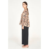 A person with long hair in a ponytail wears the Mirth Petaluma Blouse in Sand Labyrinth along with black wide-leg pants. They stand in profile against a plain background, looking forward, with arms relaxed at their sides. Their ensemble is completed by beige flat shoes, crafted from a cotton/silk blend made in India.