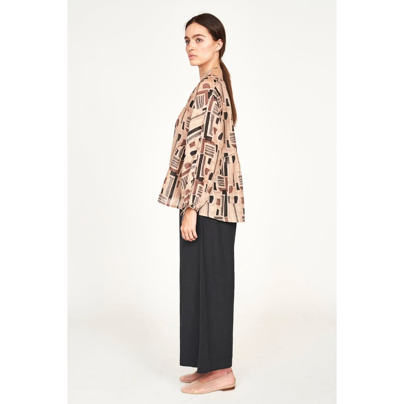 A person with long hair in a ponytail wears the Mirth Petaluma Blouse in Sand Labyrinth along with black wide-leg pants. They stand in profile against a plain background, looking forward, with arms relaxed at their sides. Their ensemble is completed by beige flat shoes, crafted from a cotton/silk blend made in India.