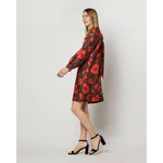 A person with long hair is walking while wearing a knee-length, long-sleeve Maggie Dress by Ann Mashburn, made from Liberty Fabrics silk and adorned with a red floral pattern on a dark background. They are also wearing black, strappy, heeled sandals. The background is a plain, light-colored backdrop.