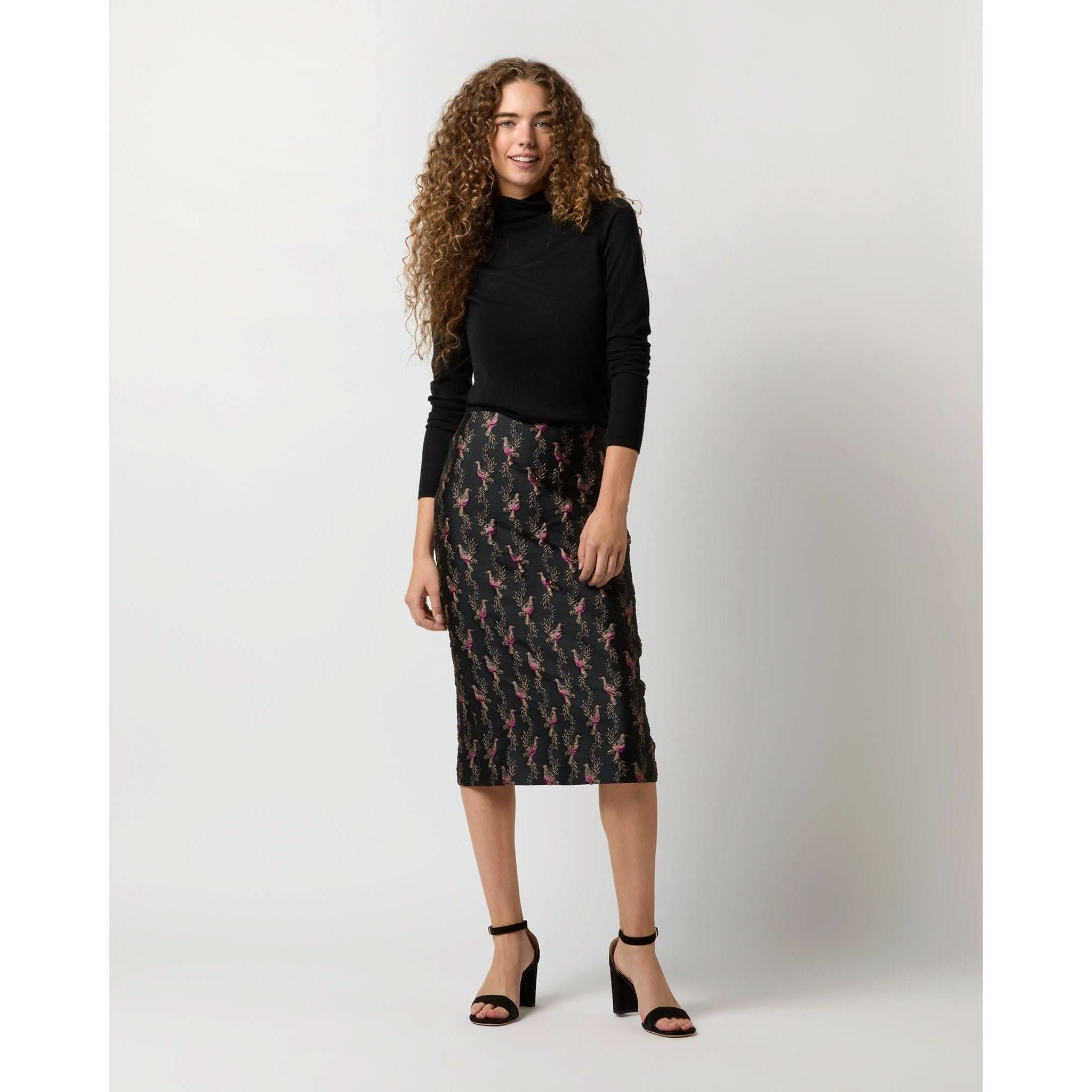 A woman with curly hair stands against a plain background, dressed in a black turtleneck and the Ann Mashburn Aurora Skirt in Black Magenta Hummingbird Matelassé, paired with black high-heeled sandals. She smiles gracefully, one hand by her side and the other slightly raised, exuding effortless elegance.
