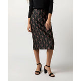 A person wearing a long-sleeved black top and an Ann Mashburn Aurora Skirt in Black Magenta Hummingbird Matelassé stands against a plain backdrop, paired with black high-heeled sandals. Their face is outside the frame in the image.