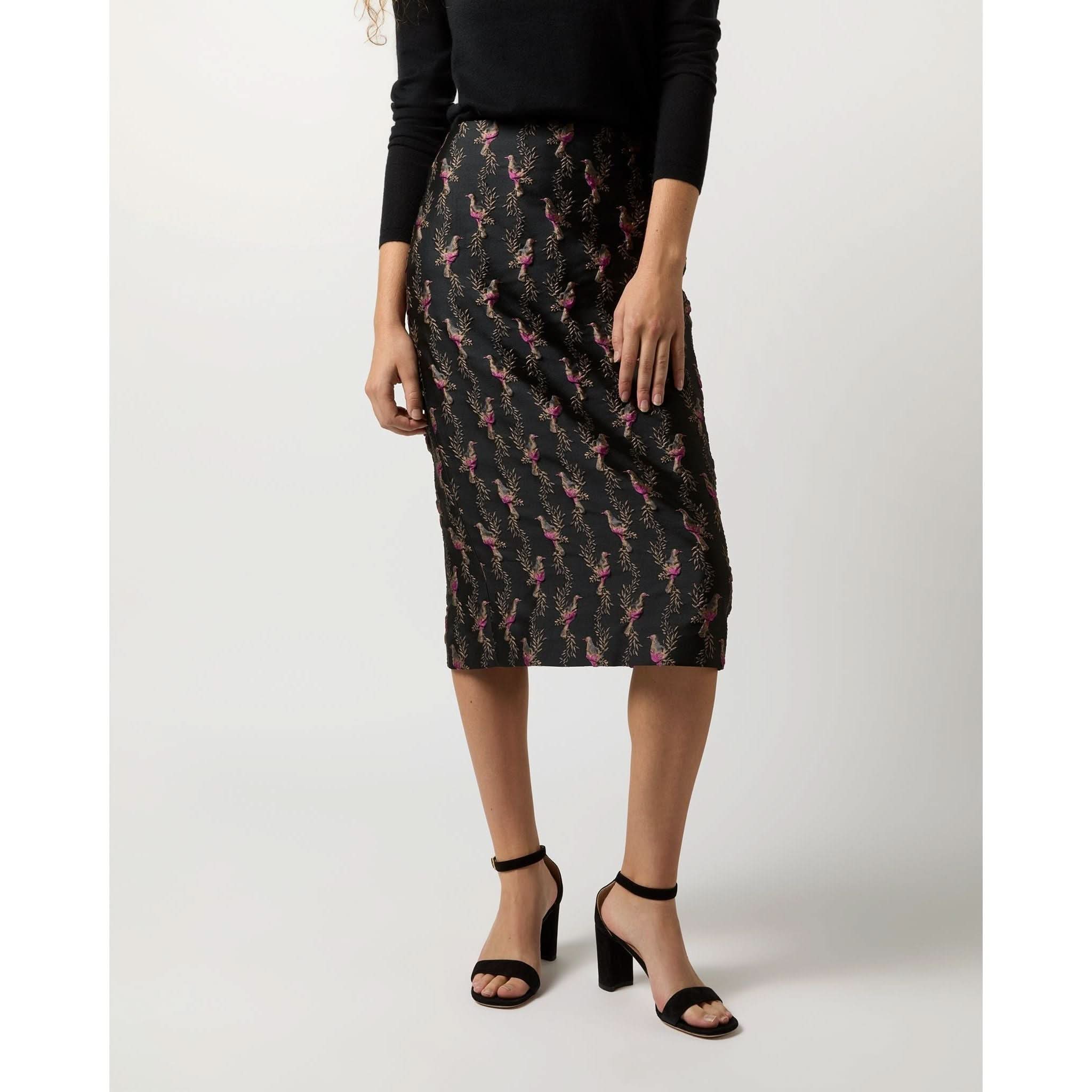 A person wearing a long-sleeved black top and an Ann Mashburn Aurora Skirt in Black Magenta Hummingbird Matelassé stands against a plain backdrop, paired with black high-heeled sandals. Their face is outside the frame in the image.
