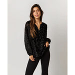 A woman with long brown hair stands against a plain background, wearing an Ann Mashburn Ariana Blouse in Black Leopard Fil Coupé Georgette and black pants. She has one hand in her pocket and displays a confident expression.