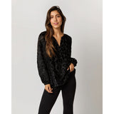 A woman with long brown hair stands against a plain background, wearing an Ann Mashburn Ariana Blouse in Black Leopard Fil Coupé Georgette and black pants. She has one hand in her pocket and displays a confident expression.