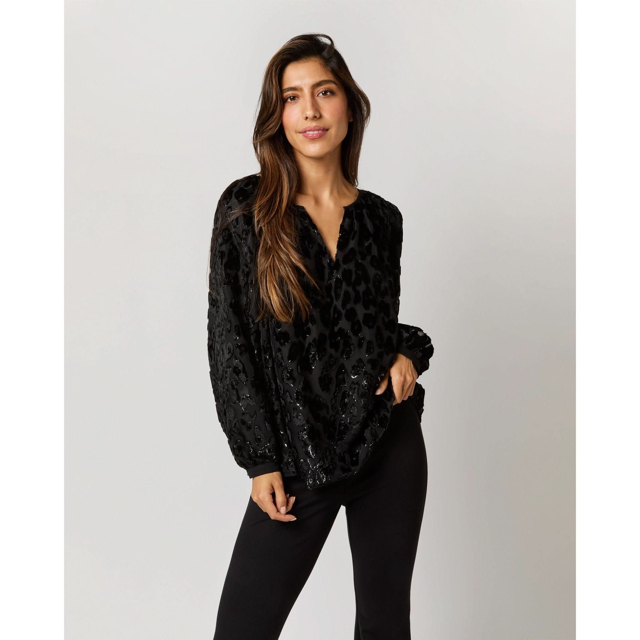 A woman with long brown hair stands against a plain background, wearing an Ann Mashburn Ariana Blouse in Black Leopard Fil Coupé Georgette and black pants. She has one hand in her pocket and displays a confident expression.