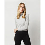 A person with long hair is standing against a plain background, wearing the Ann Mashburn Superfine Funnel-Neck Sweater in silver cashmere and black pants, effortlessly showcasing it as a versatile layering piece.