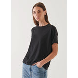 A person with long brown hair, wearing an oversized fit black Patrick Assaraf SS Iconic Boyfriend Crew T-shirt made of soft pima cotton and light blue jeans, stands against a plain light background with one hand in a pocket, looking towards the camera with a neutral expression.