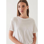 A woman with long brown hair stands against a plain light-colored background, wearing an oversized fit, Patrick Assaraf SS Iconic Boyfriend Crew White made of soft pima cotton. She has a calm expression and is posing with one hand resting at her side.