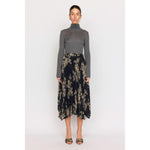 A woman in a gray turtleneck sweater and the Christy Lynn Gwen Skirt -Black Rose, featuring intricate pleating and a floral pattern, stands against a plain white background wearing open-toed heels.