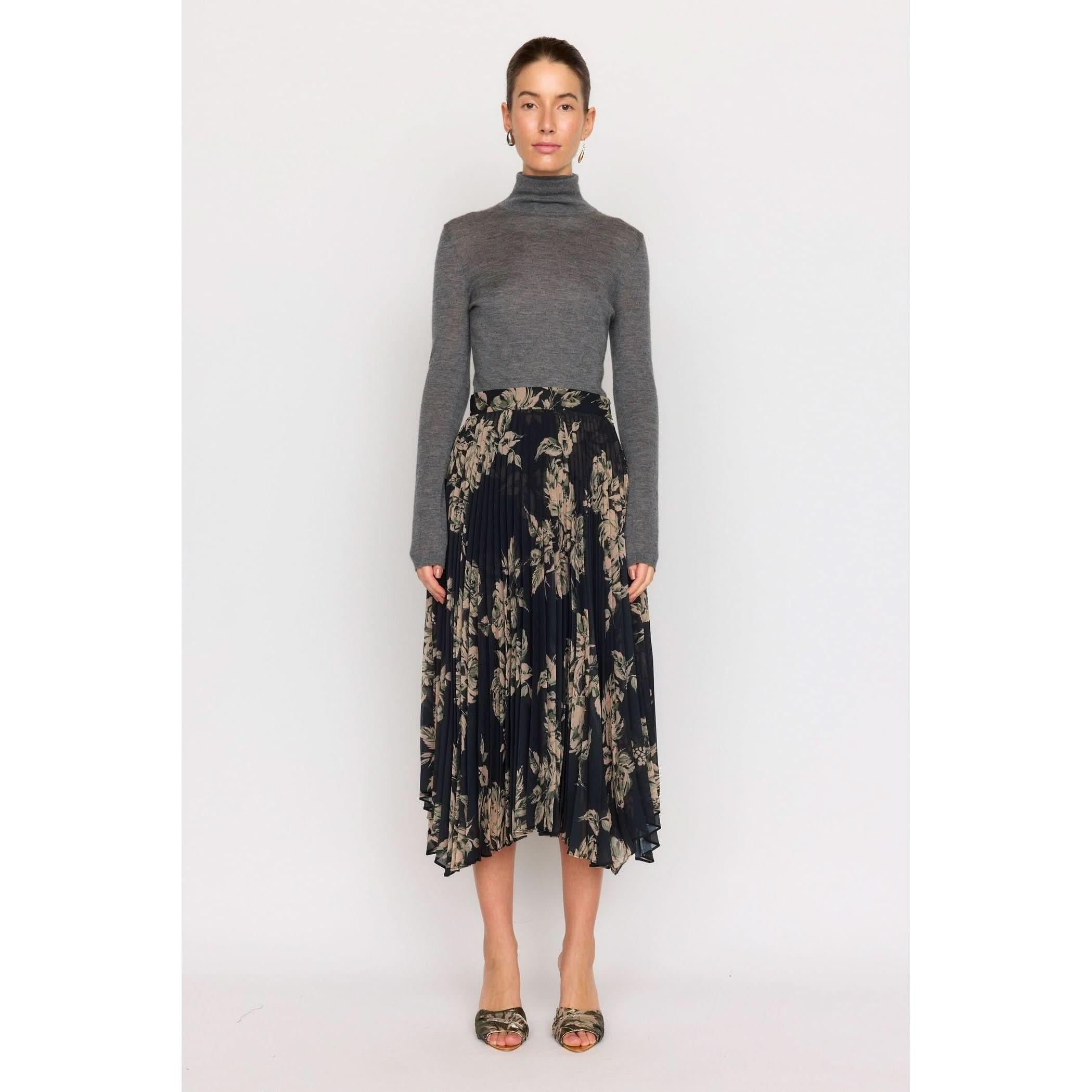 A woman in a gray turtleneck sweater and the Christy Lynn Gwen Skirt -Black Rose, featuring intricate pleating and a floral pattern, stands against a plain white background wearing open-toed heels.