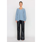 A woman stands against a plain background wearing a dusty blue Christy Lynn Allie Sweater and loose black leather pants. She has short hair and small earrings as accessories. Her expression is neutral as she faces the camera, perfectly balancing bold style with understated elegance.
