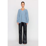 A woman stands against a plain background wearing a dusty blue Christy Lynn Allie Sweater and loose black leather pants. She has short hair and small earrings as accessories. Her expression is neutral as she faces the camera, perfectly balancing bold style with understated elegance.