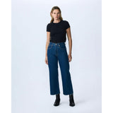 A person with shoulder-length hair stands against a plain white background. They are wearing a black t-shirt tucked into SLVRLAKE's Slvrlake Denim Grace Crop Revolution high-rise, dark blue jeans with a cropped wide leg that stops above the ankles. They are also wearing black ankle boots.
