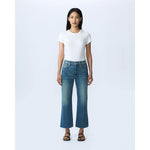 A woman stands against a plain light-colored background. She is wearing a fitted white T-shirt, SLVRLAKE's Grace Crop Blue Bird high-rise blue jeans with a slight flare and raw hem, and black sandals. She has long straight hair and a neutral expression, with her arms relaxed by her sides. The look reflects effortless style made in the U.S.A.